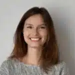 Eine Junge Frau Mit Glattem Braunem Haar Und Grauem Pullover Steht Mit Einem Strahlenden Lächeln Vor Einem Schlichten Hellen Hintergrund Und Ist Bereit, Erstklassige Reinigungsdienste Anzubieten.